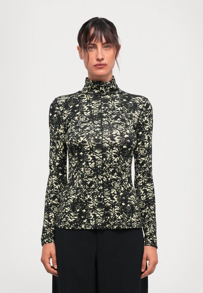 Femme aux cheveux bruns, portant un haut noir à col haut et manches longues avec un motif floral blanc, se tenant devant un fond clair et uni.
