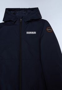 Veste bleu marine avec fermeture éclair à l'avant, capuche élastique et un patch circulaire sur la manche gauche. Tissu lisse avec une finition mate.