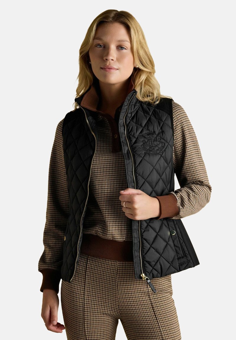 Blondynka w brązowym swetrze w zygzaki i spodniach w tym samym wzorze z brązowymi mankietami oraz pikowanej czarnej kamizelce bez rękawów, stojąca na tle białym.