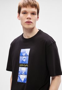 Μαύρο βαμβακερό t-shirt με κατακόρυφο σχέδιο εικονογραφημένων χαρακτήρων σε μπλε αποχρώσεις, με το κείμενο "Jujutsu Kaisen" σε κίτρινο στο κέντρο.