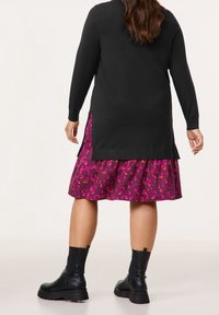 Cardigan en maille noire à manches longues, avec des fentes latérales. En dessous, une jupe rose à motifs avec un bas évasé et une texture lisse. Bottines noires.