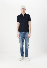 Tricou polo bleumarin cu detalii albe la guler, blugi deschiși deteriorați, și adidași albi. Modelul poartă o șapcă bej, stând pe un fundal alb.