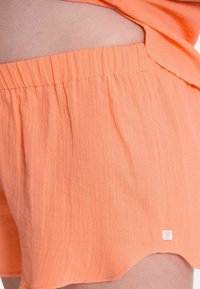 Kurze, orangefarbene Stoffshorts mit einem elastischen Bund und einem dezenten Logoshirn am Saum. Die Textur wirkt weich und leicht zerknittert.