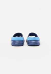 Zoccoli di gomma blu con fascia posteriore e superficie liscia. Presentano un'etichetta azzurro chiaro con "Calvin Klein Jeans" sul tallone.
