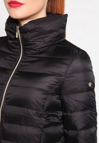Doudoune noire avec un col montant, dotée d'une texture matelassée et d'une fermeture éclair métallique. Un petit accent doré est visible sur la manche.