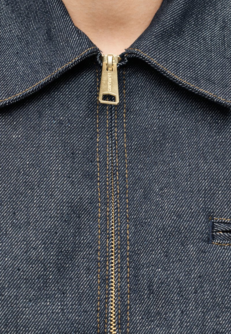 Veste en denim avec un col structuré, une fermeture éclair dorée avec un tirette à logo et des surpiqûres jaunes contrastées. Tissu bleu foncé avec une texture subtile.
