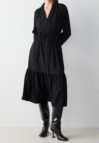 Nainen, joka on pukeutunut mustaan pitkähihaiseen midi-mekkoon, jossa on kiristettävä vyötärö, ja mustiin patenttipintaisiin polvimittaisiin korkokenkiin, seisoo valkoista seinää vasten.