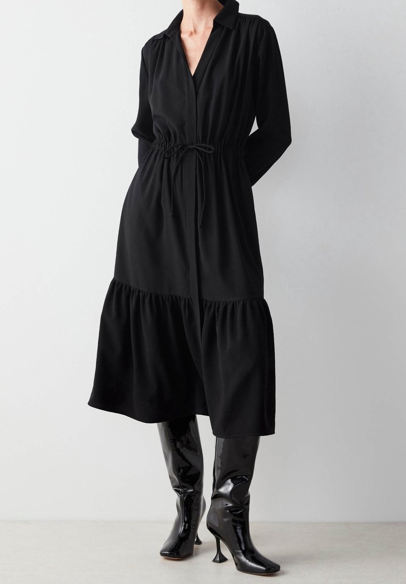 Nainen, joka on pukeutunut mustaan pitkähihaiseen midi-mekkoon, jossa on kiristettävä vyötärö, ja mustiin patenttipintaisiin polvimittaisiin korkokenkiin, seisoo valkoista seinää vasten.