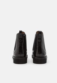 Paired de bottines Chelsea en cuir noir avec des détails brogue, des languettes de tirage et des semelles en caoutchouc noir, vues de dos sur un fond blanc.
