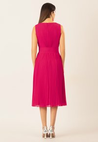 Abito plissettato fucsia con vita aderente e design senza maniche. Presenta un collo rotondo e una cerniera nascosta sul retro. Scarpe con tacco alto argentate.