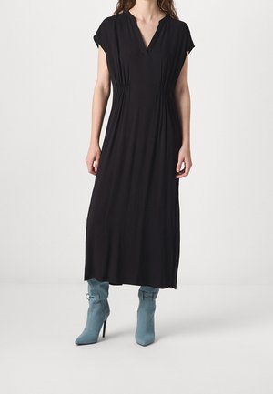 Femme portant une robe midi noire avec un col en V et une taille froncée, associée à des bottines à talons hauts bleu clair sur un fond uni.