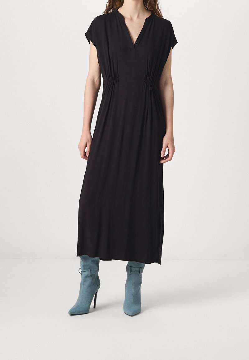 Femme portant une robe midi noire avec un col en V et une taille froncée, associée à des bottines à talons hauts bleu clair sur un fond uni.