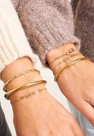 Twee polsen versierd met verschillende gouden armbanden: een gladde bangle, een kralenbangle en een bedelarmband met de tekst "BESTIES." De texturen variëren.