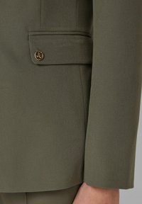 Blazer verde oliva con un acabado texturizado, que cuenta con un bolsillo lateral y herrajes de botones en tono dorado. Corte ajustado con hombros estructurados.