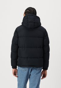 Sort polstret jakke med en stor hætte, kviltet design, elastiske manchetter og en afslappet pasform, parret med lyseblå denimjeans.