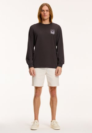 Long-sleeve dark gray T-shirt with a small white graphic design, paired with light beige shorts and cream-colored sneakers.