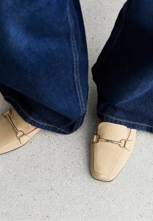Beige ruskind loafers med metalstangdetalje båret med vidbenede, mørkeblå cowboybukser på en grågulvet, der er plettet.