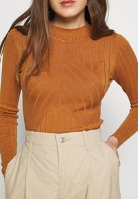 Rostorange gerippter Pullover mit hohem Kragen und Zickzackmuster, kombiniert mit hellbeigen, hoch taillierten Hosen mit seitlichen Taschen.