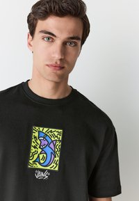 Black cotton t-shirt featuring a graphic print of a blue shark on a green background with wavy lines, and "JAWS" text at the bottom.