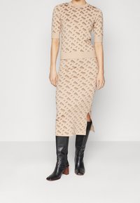 Top beige a maglia e gonna midi con motivo a diamante in rilievo. La gonna presenta uno spacco laterale. Abbinata a stivali neri alti fino al ginocchio con tacco.