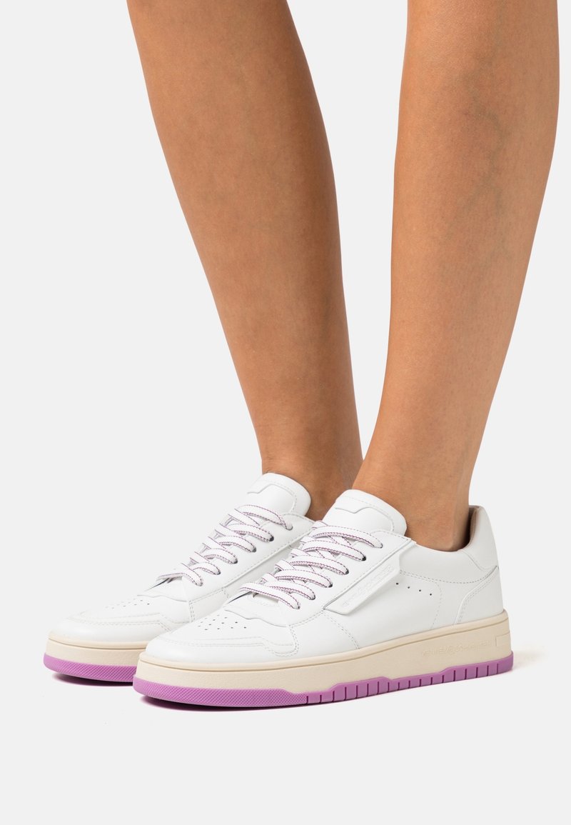 Baskets basses blanches avec semelles violettes et lacets violets assortis portées sur des jambes nues, sur un fond uni.