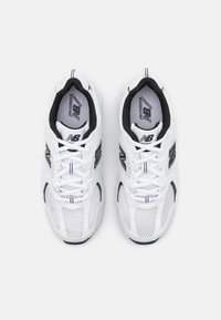 Chaussures de course New Balance blanches avec des accents noirs, en tissu mesh, avec des lacets, vues de dessus sur un fond gris clair.