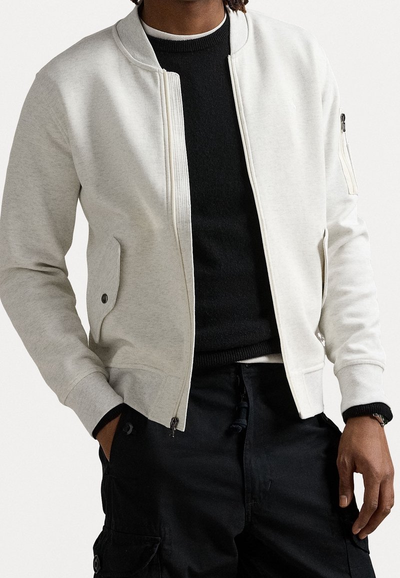Homme portant une veste bomber gris clair sur un pull noir avec un pantalon cargo foncé, mains partiellement dans les poches, sur un fond uni.