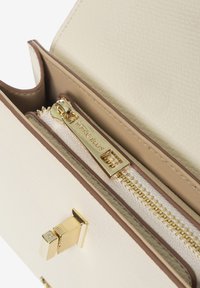 Creme farbene Kunstledertasche mit goldenen Akzenten, ausgestattet mit einer Reißverschlusstasche, beiger Innenauskleidung und einem dekorativen Verschlussdetail.