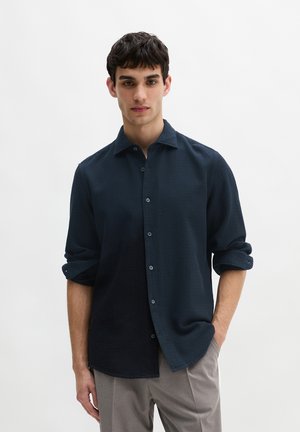 Dunkelblaues, strukturiertes Hemd mit knöpfbarem Kragen und langen, leicht aufgekrempelten Ärmeln. Vorderknöpfe und lockere Passform. Kombiniert mit hellen Hosen.