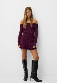Purple off-the-shoulder dress with ruched detailing, long sleeves, and layered skirt, paired with black knee-high stiletto boots.