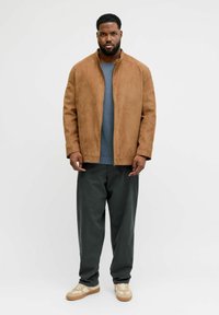 Brun semsket jakke med oppreist krage, kombinert med en blå genser og mørkegrønne vide bukser, fullført med beige og kremfargede joggesko.