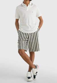 Person som bär en vit polo-shirt, svart och vit vertikalt randig shorts, vita sneakers med svarta och röda accenter, stående med benen i kors.