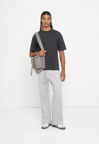Siva oversized majica, svijetlosive trenirke i siva torba preko tijela. Model nosi crno-bijele tenisice i stoji protiv bijele pozadine.