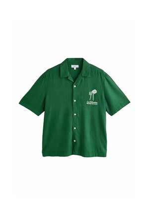 Młody mężczyzna stoi obok basenu, ubrany w zieloną koszulę z krótkim rękawem i pasujące krótkie spodenki, beżowe buty, noszący beżową torbę na jednym ramieniu.