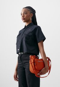 Black cropped shirt with two front pockets, paired with dark pants. Orange bag with multiple compartments and a strap, held by the model.