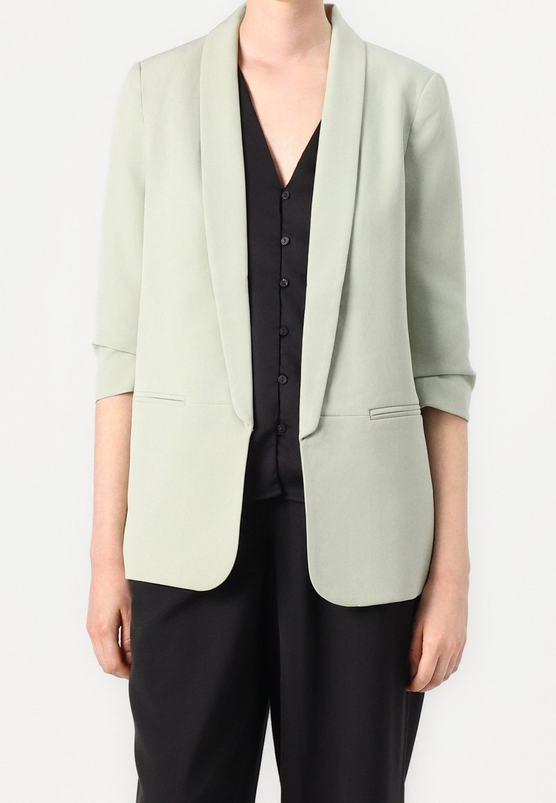 Personne portant un blazer vert clair avec les manches retroussées sur une chemise noire boutonnée et un pantalon noir, visible du cou jusqu'à mi-cuisse.