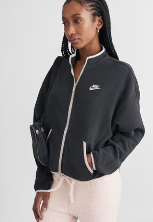 Čierna fleecová bunda na zips s bielymi akcentmi a logom Nike. Model má na sebe svetloružové, huňaté nohavice s šnúrkami. Minimalistický dizajn.