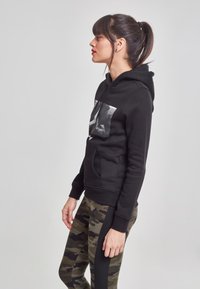 Sudadera negra con bolsillo delantero, que presenta una gran impresión gráfica. Combinada con leggins de camuflaje, que exhiben un tejido texturizado y un diseño ajustado.