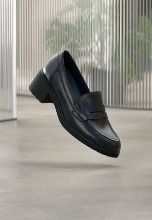 Mocasín de cuero negro con tacón grueso que flota sobre un suelo de hormigón liso, con persianas verticales borrosas y plantas en el fondo.