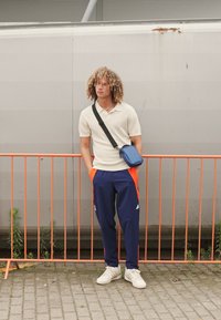 Polo shirt bianco a righe, pantaloni blu con strisce arancioni, sneakers bianche e una borsa a tracolla blu. Lo sfondo presenta una barriera arancione.