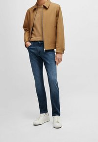 Béžová zipová bunda, béžové tričko, tmavě modré džíny, bílé tenisky. Bunda má límec a boční kapsy. Neformální outfit s volným střihem.