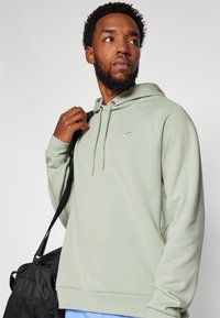 Lys grønn hettegenser med snorhette, raglanermer og en liten svart Nike-logo på brystet. Brukt med en svart veske.