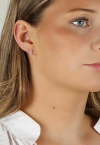 Silverfärgade hoopsmycken med en rad av små klara ädelstenar längs den främre kanten, placerade mot en neutral hudton.