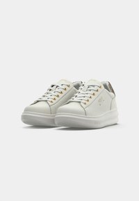 Lave hvite sneakers med gullhull, hvite snørebånd og en slangeskinn-mønstrede hæltab angitt mot en hvit bakgrunn.