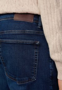 Mørkeblå jeans med en glat tekstur, prydet med brun læderbranding ved taljen og orange syninger på lommerne.