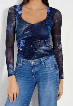 Kvinde iført tætsiddende, gennemsigtig navyblå langærmet top med blomsterprint og højtaljede lyseblå jeans.