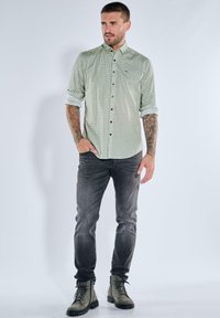 Mann mit hochgekrempelten Ärmeln, der ein gemustertes hellgrünes Hemd, ausgewaschene schwarze Jeans und graue Schnürstiefel trägt, vor einem schlichten Hintergrund stehend.