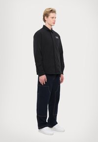 Svart skjorta med knäppning och en subtil logotyp, tillsammans med marinblå byxor med orange sömnadsdetaljer och vita sneakers.