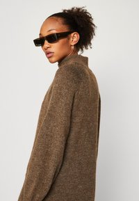Brązowy sweter dzianinowy z wysokim kołnierzem, charakteryzujący się teksturowanym wykończeniem. Model nosi ciemne okulary przeciwsłoneczne i ma kręcone włosy.