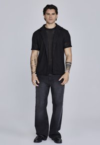 Joven con cabello oscuro que está de pie vistiendo una camisa corta de manga corta negra y transparente, un cárdigan negro de punto abierto, jeans oscuros de pierna ancha y zapatos negros.